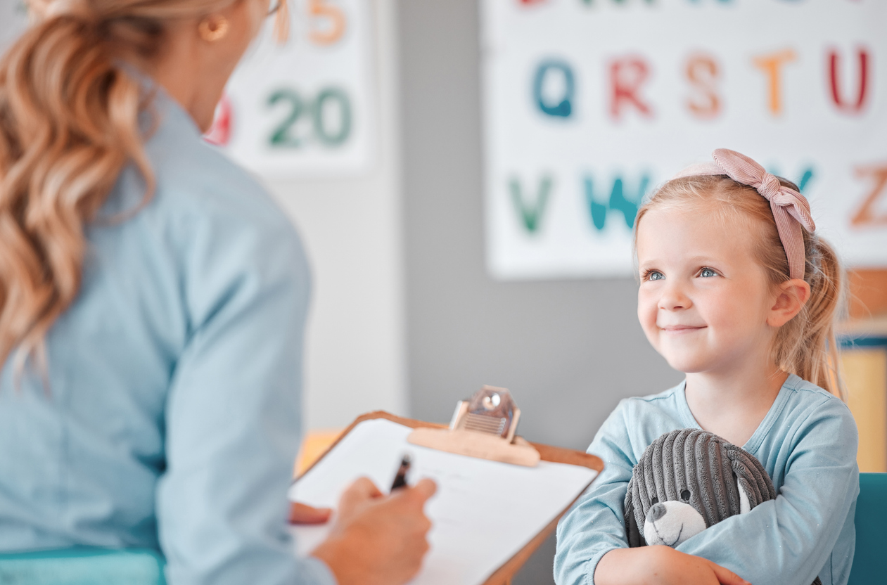 Young smiling caucasian child sitting with her psychologist in a clinic. Cute girl talking to a mental health professional. Using a clipboard to take notes on her patient. Dyslexia and anxiety